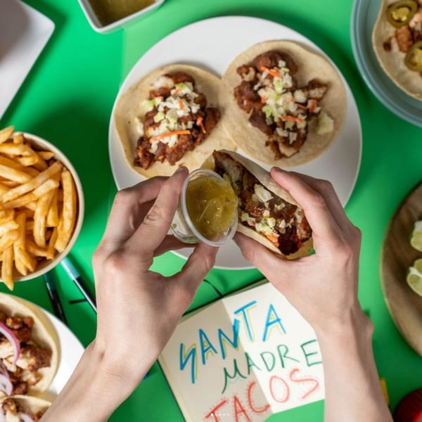 Hands squeezing green salsa onto a Santa Madre Taco Shop taco, with fries and other tacos nearby.