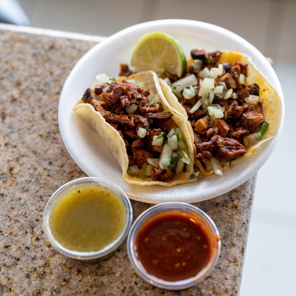 Two Santa Madre Taco Shop tacos with meat, onions, cilantro, lime, and two salsa cups.