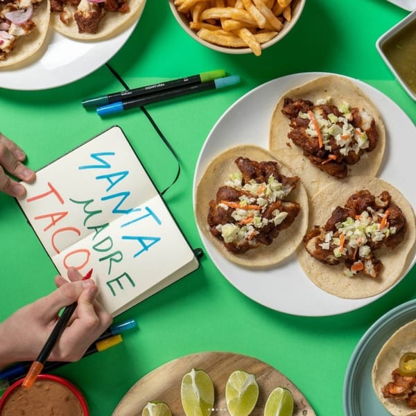 Overhead view of Santa Madre Taco Shop tacos, fries, and a person writing "Santa Madre Taco" in a notebook.