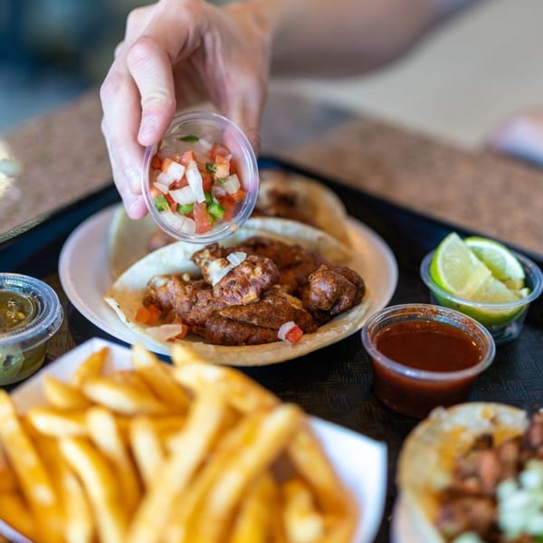 Hand adding pico de gallo to a Santa Madre Taco Shop taco, with fries and sauces.