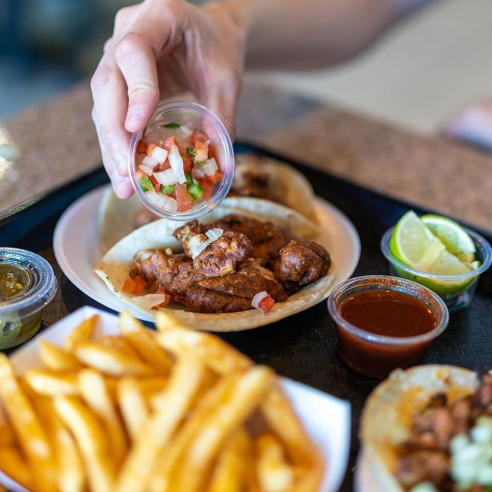 Hand adding pico de gallo to a Santa Madre Taco Shop taco, with fries and sauces.